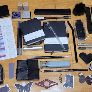 May include: An assortment of stationery and accessories, including black pocket notebooks, pens, stickers, and a reading log. The items are arranged on a wooden surface, with a variety of shapes and sizes.