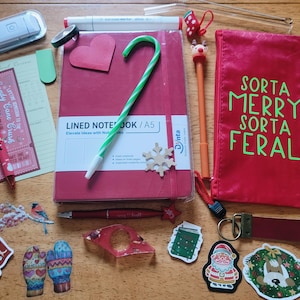 May include: A red A5 lined notebook with a green and white candy cane pen, a red pencil case with the text SORTA MERRY SORTA FERAL, various holiday-themed stickers, a red pen, a green marker, a red marker, a bookmark, and a keychain.