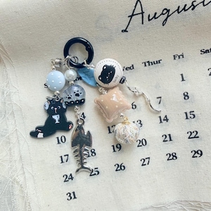 May include: A keychain featuring a black ring with multiple charms including a black cat figure, a silver fish skeleton, a blue leaf-shaped tag with the word "Lucky", a white bead with a cat face, a beige pillow charm, and a textured sphere.
