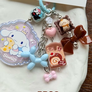 May include: A collection of colorful charms on a light blue clasp. Charms include a cartoon dog, a pink heart, a blue balloon dog, a bow, and a Sanrio character. The text "Sweet Cookie" and "Keep Luck" are visible on one charm. The code "BB03" is also visible.