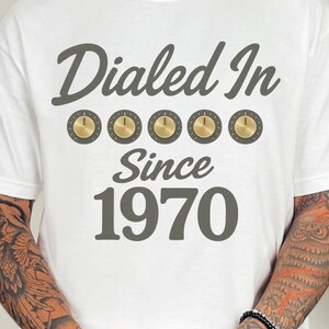 May include: White t-shirt with the text "Dialed In Since 1970" in gray script. Above the text are five gold-colored knobs. The shirt is a casual style, suitable for everyday wear.