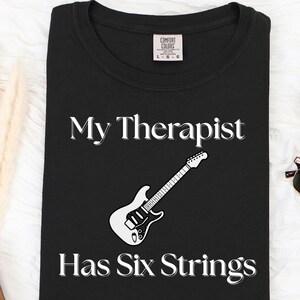 May include: A black short-sleeved t-shirt featuring the white text My Therapist Has Six Strings and a graphic of a white electric guitar. The shirt has a Comfort Colors brand label at the inner neckline indicating a size large.