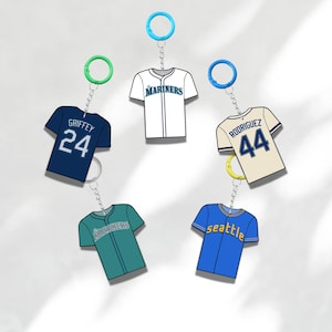 May include: Keychains featuring miniature baseball jerseys in various colors, including navy blue, teal, and white. Each keychain has a different player's name and number, with a blue, green, or yellow ring.
