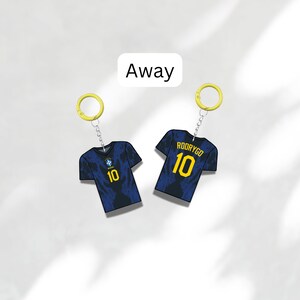 Puede incluir: Dos llaveros con camisetas de f&uacute;tbol en miniatura. Las camisetas son de color azul oscuro con detalles amarillos, incluido el n&uacute;mero 10. Un llavero tiene el nombre "RODRYGO" en la espalda. La palabra "Away" est&aacute; en un cuadro blanco arriba.