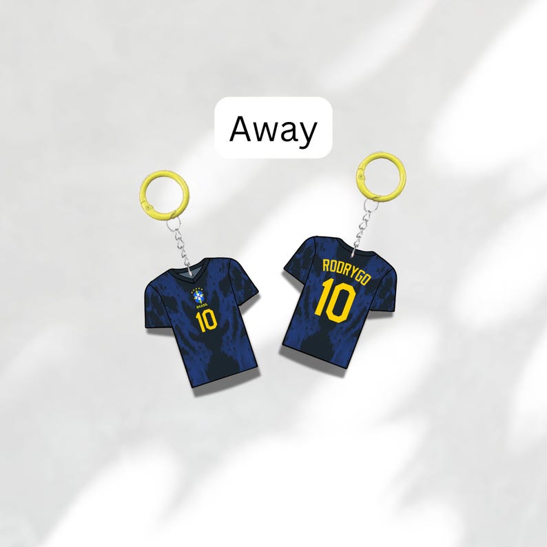 Puede incluir: Dos llaveros con camisetas de f&uacute;tbol en miniatura. Las camisetas son de color azul oscuro con detalles amarillos, incluido el n&uacute;mero 10. Un llavero tiene el nombre "RODRYGO" en la espalda. La palabra "Away" est&aacute; en un cuadro blanco arriba.