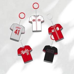 May include: A collection of baseball jersey keychains in various colors, including red, white, gray, and black. Each keychain features a jersey design with numbers and team names, attached to a metal chain and a red or white ring.