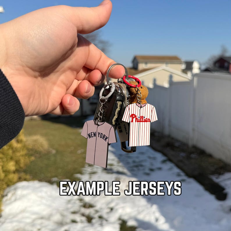 Puede incluir: Un llavero con dos colgantes en miniatura de camisetas de b&eacute;isbol. Una es una camiseta rosa claro con "NEW YORK" impreso, y la otra es una camiseta a rayas blancas y rojas con "Phillies" impreso. El texto "EXAMPLE JERSEYS" est&aacute; en la parte inferior.