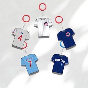 Könnte beinhalten: Baseball-Trikot-Schlüsselanhänger in verschiedenen Farben, darunter Grau, Weiß, Blau und Marineblau. Jeder Schlüsselanhänger zeigt ein Miniatur-Trikot-Design mit Teamlogos, Spielernamen und Nummern. Die Schlüsselanhänger sind an roten oder blauen Schlüsselringen befestigt.