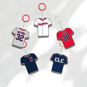Puede incluir: Una colección de llaveros con camisetas de béisbol en varios colores, incluyendo rojo, blanco, azul marino y gris. Cada llavero presenta un diseño de camiseta con nombres y números de jugadores, y logotipos de equipos.