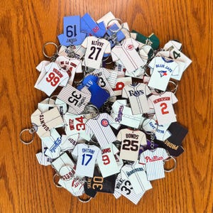 May include: A collection of baseball jersey-shaped keychains in various team colors. Each keychain features a player's name and number, with team logos and details. The keychains are attached to silver keyrings and are arranged on a wooden surface.