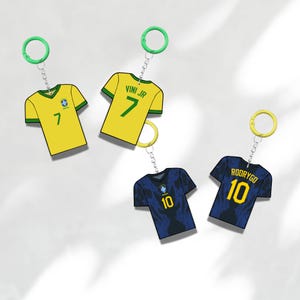 Puede incluir: Tres llaveros de camisetas de f&uacute;tbol. Dos son amarillas con ribetes verdes, una con el n&uacute;mero 7 y la otra con "VINI JR" y el n&uacute;mero 7. Las otras dos son azul marino con ribetes amarillos y el n&uacute;mero 10.