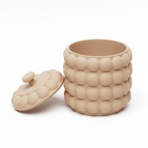 Puede incluir: Un recipiente beige con tapa a juego. El recipiente y la tapa están decorados con un patrón repetitivo de formas redondeadas. La tapa está a un lado, revelando el interior del recipiente. El recipiente está sobre un fondo blanco.