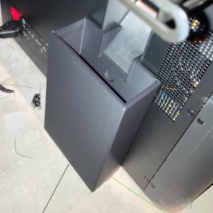 Könnte beinhalten: Ein dunkelgraues, rechteckiges 3D-gedrucktes Objekt mit offener Oberseite und Stufendesign. Das Objekt befindet sich neben einer schwarzen Computerkomponente mit einem Mesh-Lüfter.