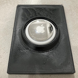 Puede incluir: Soporte negro impreso en 3D para un AirTag de Apple. El soporte tiene forma rectangular con una superficie texturizada y un hueco circular para sujetar el AirTag. El AirTag es plateado con el logotipo de Apple.