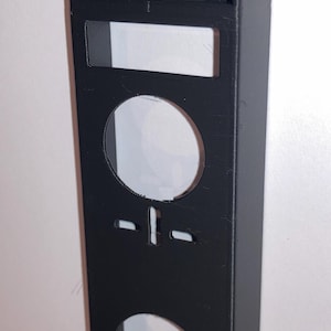 May include: A black, rectangular, 3D-printed object with a box-like top and a vertical panel. The panel has two circular cutouts and a rectangular cutout. The object is mounted on a white surface.