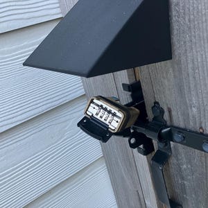 May include: A combination lock with a brass-coloured frame is attached to a black metal gate latch. The lock's numbers are visible. The gate is made of weathered wood and is mounted on a white siding wall. A black awning is above the gate.