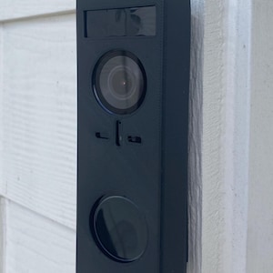 May include: A black rectangular doorbell camera mounted on a white wooden wall. The camera has a circular lens, a smaller circular button, and a rectangular window at the top. The doorbell is a sleek, modern design.