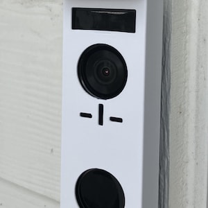 May include: A white rectangular video doorbell featuring a top solar panel, a central camera lens, a black horizontal and vertical line icon, and a large circular black button at the bottom. The device is mounted vertically on a white surface.