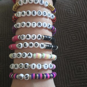 May include: A collection of beaded bracelets spelling out words like "YOUR", "GOLDEN", and "TAKEDOWN". The bracelets feature a variety of colors including pink, black, gold, and purple, with white letter beads. The bracelets are stacked on a wrist.