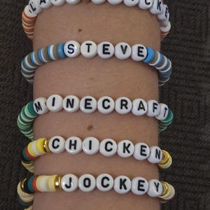 May include: Five colorful beaded bracelets spelling out "LAVA CHICKEN", "STEVE", "MINECRAFT", "CHICKEN", and "JOCKEY" in white letter beads. The bracelets are on a wrist.