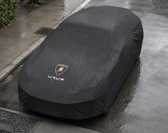 May include: A black car cover draped over a vehicle, featuring the gold Lamborghini shield logo and the white text "URUS" printed on the hood. The fabric appears damp with visible water droplets, indicating a rainy outdoor environment.