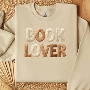 May include: A beige sweatshirt featuring the words "BOOK LOVER" in textured, 3D lettering. The letters are in shades of beige and brown. The sweatshirt is a neutral colour and has a crew neck.