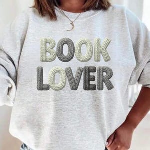 May include: A pale grey sweatshirt featuring the text "BOOK LOVER" in a 3D knitted design. The letters are in shades of white and grey, giving a textured look. The sweatshirt is a casual style.