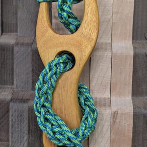May include: A light brown wooden slingshot with a Y-shaped frame featuring a central circular hole and a bottom hole. A braided blue and green paracord is threaded through the holes and tied in a knot at the top of the wooden frame prongs.