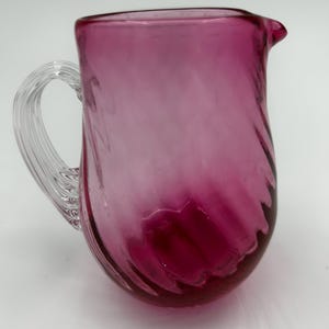 May include: A magenta-coloured glass jug with a clear, looped handle and a pouring spout. The jug has a swirled design throughout the body, creating a textured appearance. The glass is translucent, allowing light to pass through.