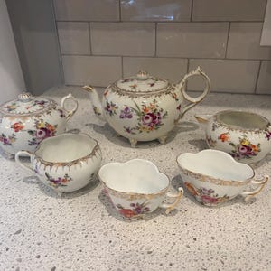 Può includere: Un servizio da tè in porcellana vintage con disegni floreali. Il set comprende una teiera, una zuccheriera, una lattiera e due tazze da tè. I pezzi sono bianchi con finiture dorate e fiori colorati.