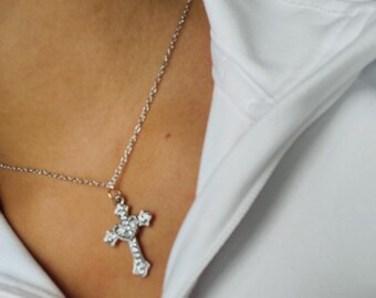 May include: A silver-colored cross pendant necklace with clear, sparkling stones. The cross is suspended from a delicate chain. The necklace is worn over a white garment.