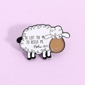 May include: A white sheep-shaped enamel pin with black accents. The pin features the text "HE LEFT THE 99 TO RESCUE ME. Matthew 18:12" in black script. The sheep has a brown face and black feet, set against a light purple background.