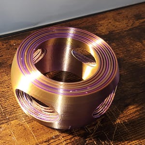 May include: A metallic, ring-shaped object with concentric circles and oval cutouts. The object has a gold and purple colour scheme, with purple accenting the edges of the circles. It sits on a wooden surface.