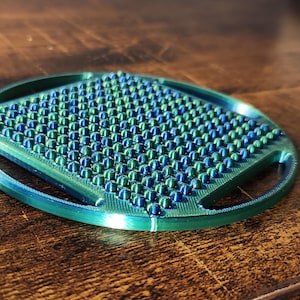 Può includere: Oggetto rotondo e strutturato con una sfumatura blu e verde. La superficie è ricoperta da piccole protuberanze arrotondate. L'oggetto ha una cornice circolare con maniglie ritagliate. Potrebbe essere uno strumento per massaggi o un elemento decorativo.