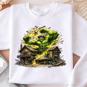 Puede incluir: Sudadera blanca con un gráfico colorido de Hulk saliendo de una casa de madera. Hulk se representa con piel verde y cabello oscuro. La casa está detallada con iluminación cálida y rodeada de escombros.