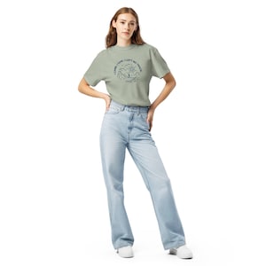 Op de afbeelding: Een vrouw draagt een saliegroen t-shirt en lichtblauwe jeans. Het t-shirt heeft een cirkelvormige afbeelding met de tekst "I CAME, I SAW, I LEFT NO TRACE" en een berg- en zonontwerp. De jeans zijn hoog in de taille en wijd uitlopend.