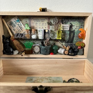 Op de afbeelding: Houten kist met een verzameling miniatuuritems met een heksenthema. Inclusief een zwart kattenbeeldje, een bezem, glazen flessen, een kikker, een kristallen bol en een rol. De kist toont ook de tekst: "MAGICAL HERBS & THEIR USES" en "The Green Witch".