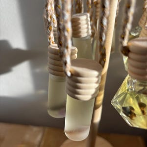 Puede incluir: Varias botellas de vidrio pequeñas con tapas de madera están suspendidas de un soporte de madera. Las botellas contienen un líquido de color claro y están unidas al soporte con cuerdas trenzadas. El soporte es de madera clara.