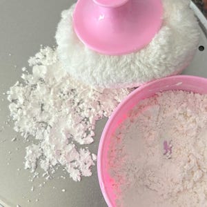 以下が含まれることがあります： 白いふわふわのパッドが付いたピンク色のパフが、白い粉末で満たされたピンク色の容器の隣にあります。白い粉末の山が銀色の表面に散らばっており、化粧品またはスキンケア製品を示唆しています。