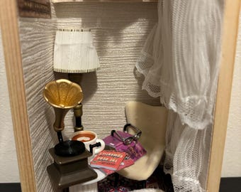 May include: Miniature diorama of a cozy reading nook. Features a small white dog on a patterned rug, a tiny armchair, and a gramophone. Bookshelves are filled with miniature books, and a lamp provides soft lighting. The Silent Patient book is visible.