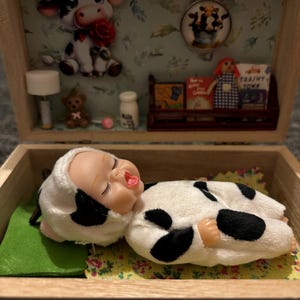 Puede incluir: Diorama en caja de madera con un muñeco de bebé disfrazado de vaca. La caja está decorada con una figura de vaca, un colgante con vacas, libros en miniatura y un osito de peluche. El muñeco de bebé está sobre una tela estampada floral.
