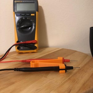 May include: A yellow and black Fluke 179 True RMS Multimeter with red and black test leads. Two test probes, one red and one black, are on a wooden surface. The multimeter has a digital display.