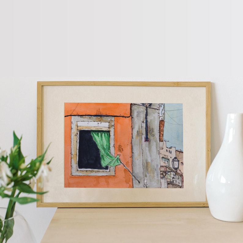May include: A framed watercolor painting depicting an orange building facade with a single window featuring a green curtain blowing in the breeze. The artwork includes architectural details of a neighboring structure and a street lamp in a soft style.