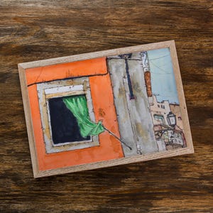 May include: A framed watercolor painting depicting an orange building facade with a dark window and a bright green curtain blowing in the breeze. The scene includes a gray wall section, a street lamp, and architectural details of nearby buildings.