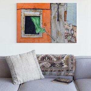 May include: A rectangular canvas wall art print featuring a watercolor painting of an orange building facade with a dark window, a green curtain blowing outward, and a street lamp. The artwork is displayed above a gray sofa with a patterned throw pillow.