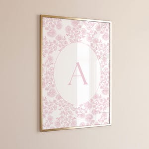 May include: A framed print with a gold-coloured frame, displaying a pink floral pattern. A large letter "A" is centred within a white oval. The floral design surrounds the oval, forming a decorative border.
