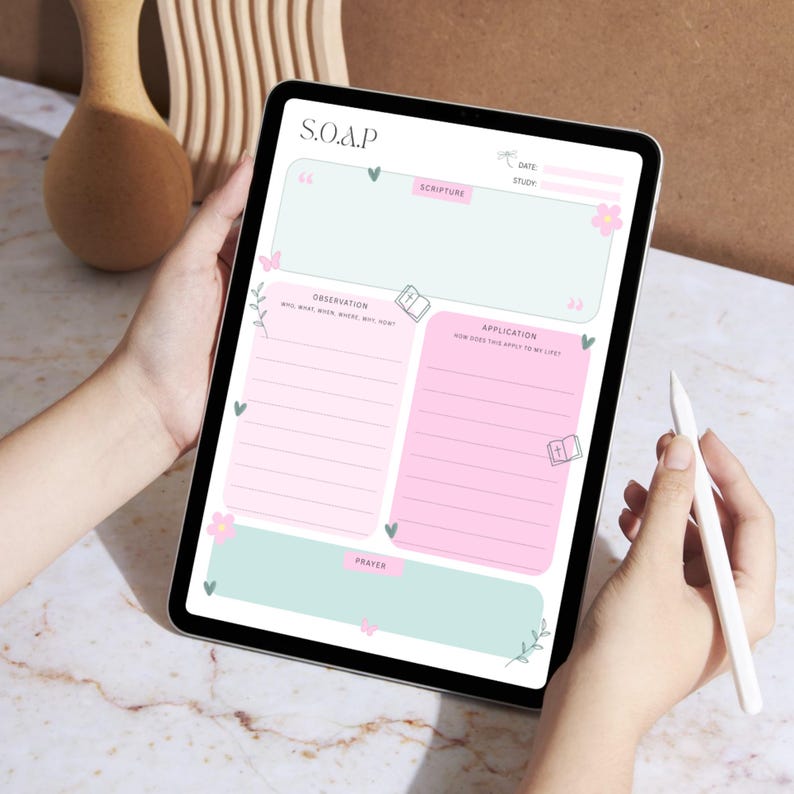 May include: A digital tablet displaying a pastel pink and mint green S.O.A.P. study template. The template includes sections for Scripture, Observation, Application, and Prayer, with floral and butterfly accents. A white stylus is held in the hand.