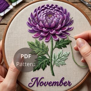 May include: A round wooden embroidery hoop containing a beige fabric panel with a hand-stitched purple chrysanthemum flower, green leaves, and the word November in purple script. A needle with green thread is positioned near the flower petals.
