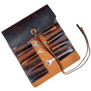 May include: A rolled brown leather tool roll with multiple pockets, holding several spanners. The leather has a distressed, vintage appearance with darker brown accents. A brass grommet and leather tie secure the roll.