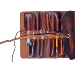 Puede incluir: Un rollo de herramientas de cuero marrón con múltiples bolsillos, que contiene varias llaves y destornilladores. El rollo se asegura con una correa de cuero y tiene un ojal de latón. El cuero tiene un acabado envejecido.